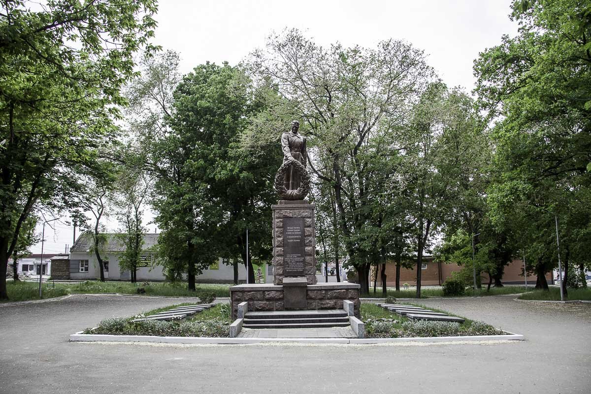 Каменный солдат с траурным венком в руке замаскировался в зелени деревьев