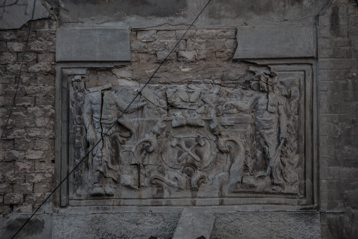 Полуразрушенный барельеф, на котором изображены два рабочих, держущих картуш с двумя скрещенными молотками